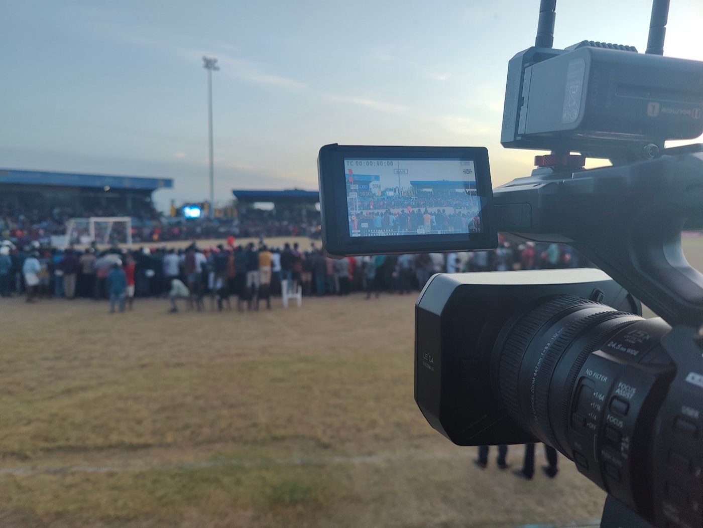 A camera on the sideline of a packed football ground at dusk